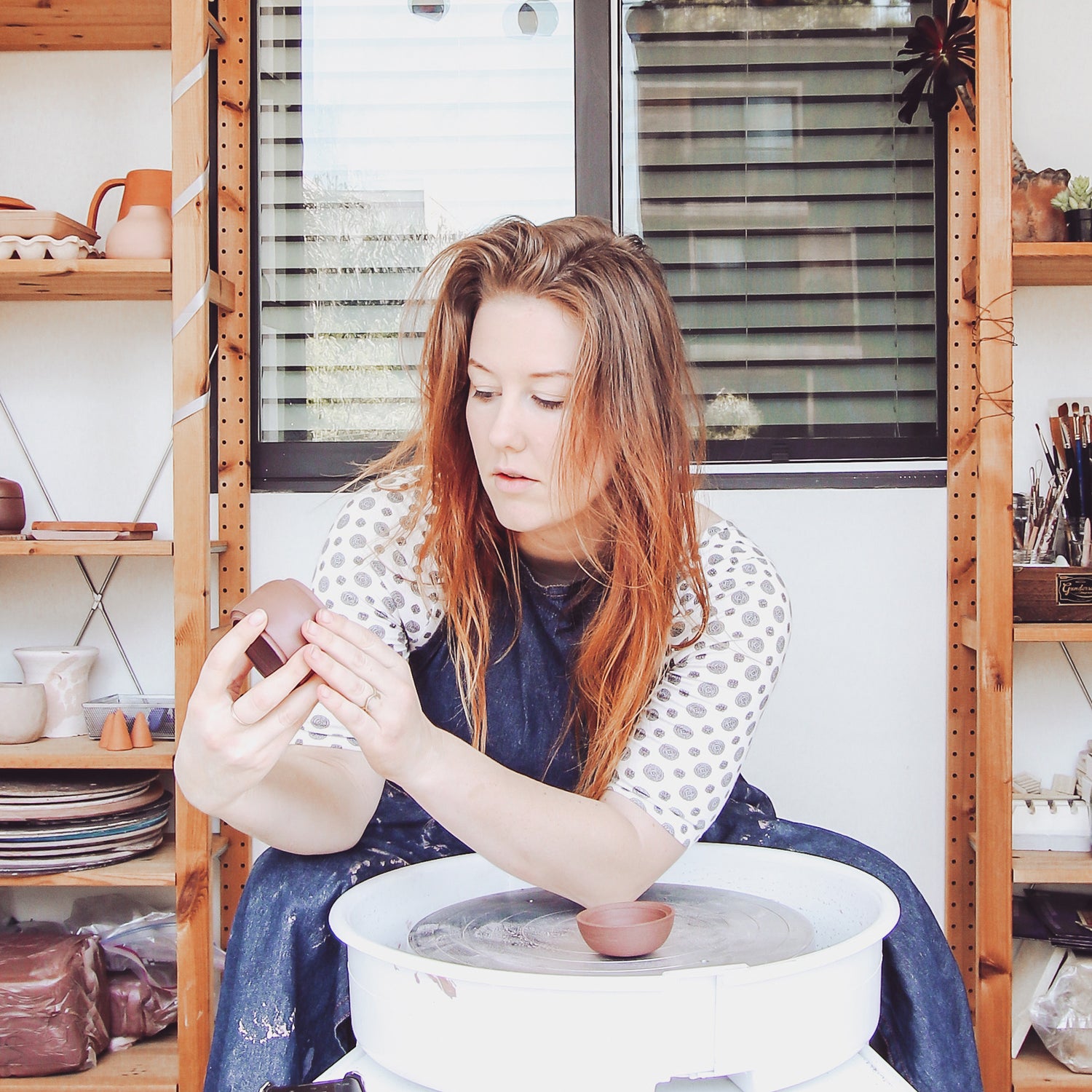female ceramic artist shaping a small clay bowl on a pottery wheel in a home studio.