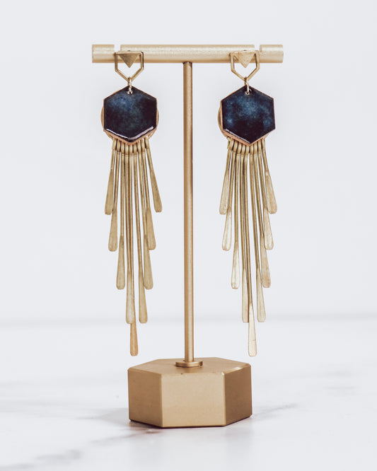 Long gold fringe tassel earrings with hexagon-shaped ceramic tops, shown in midnight blue on a gold stand.