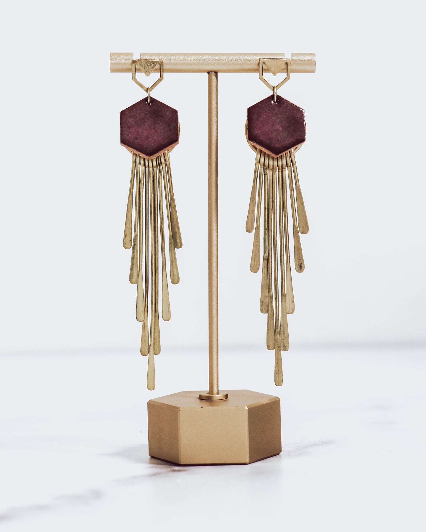 Long gold fringe tassel earrings with hexagon-shaped ceramic tops, shown in burgundy on a gold stand.