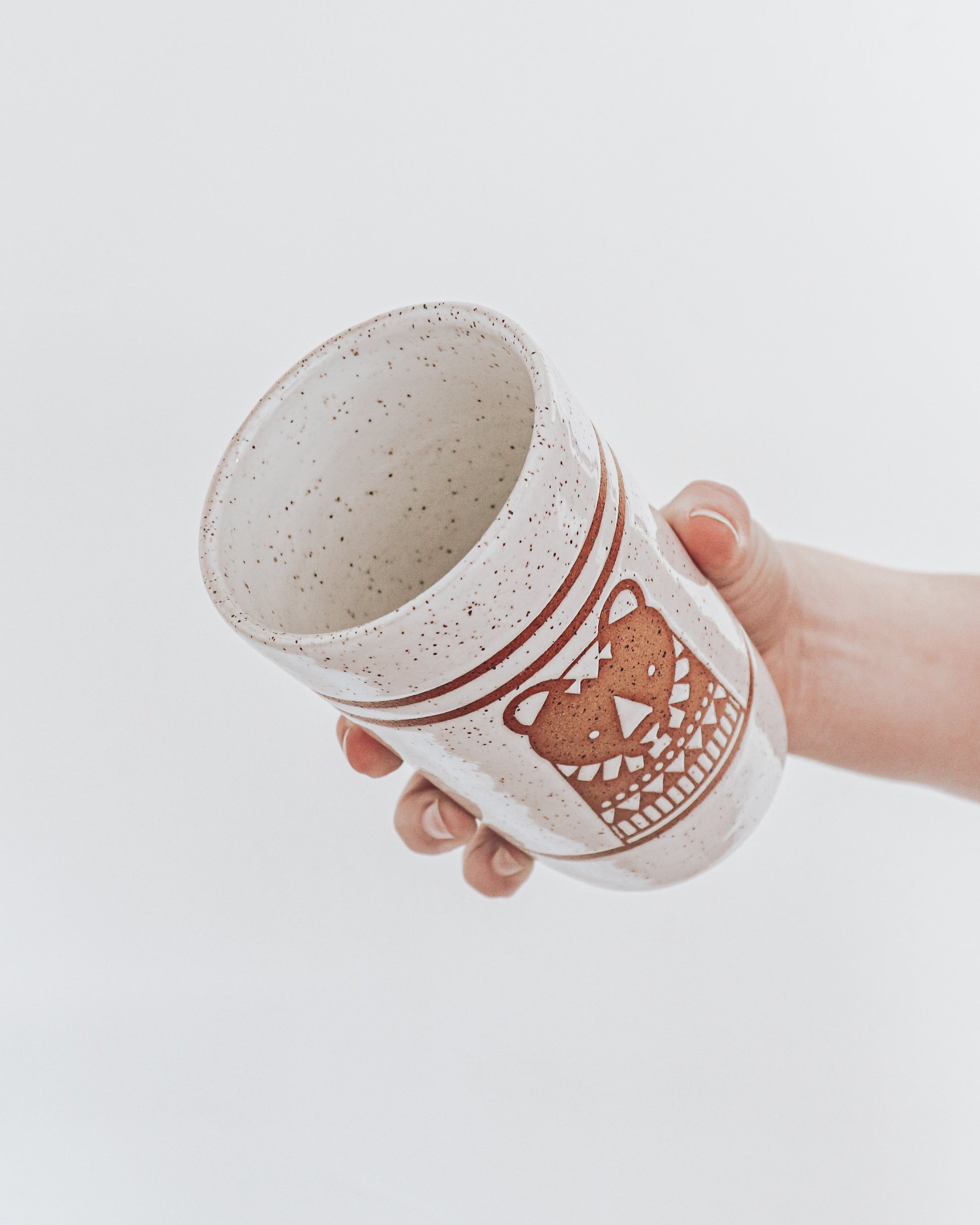 Inside view of Hand Holding a handmade speckled ceramic tumbler featuring a brown illustrated bear design with geometric detailing. 