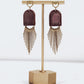 Ceramic and brass arch tassel earrings with gold fringe, shown in burgundy on a gold stand.