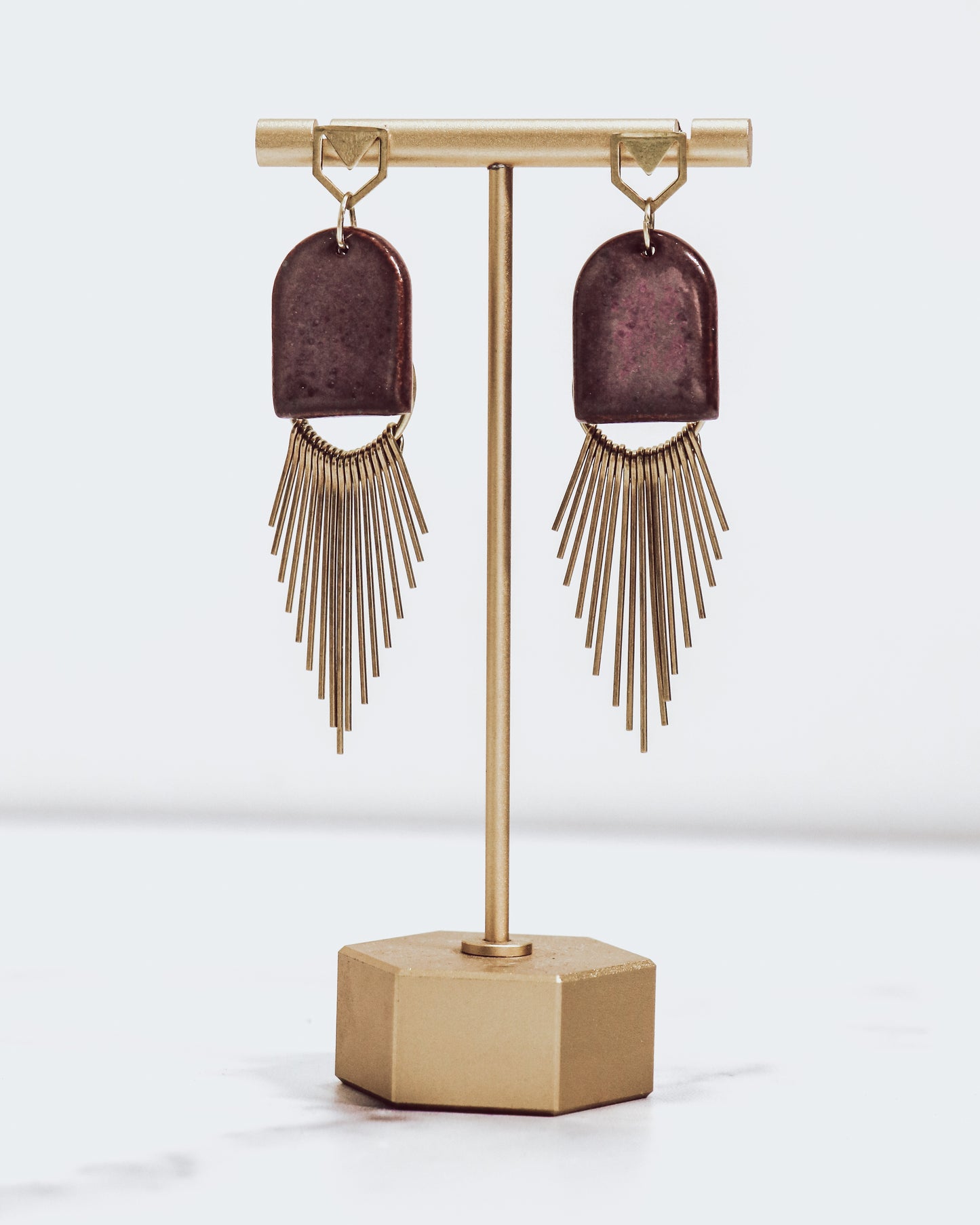 Ceramic and brass arch tassel earrings with gold fringe, shown in burgundy on a gold stand.