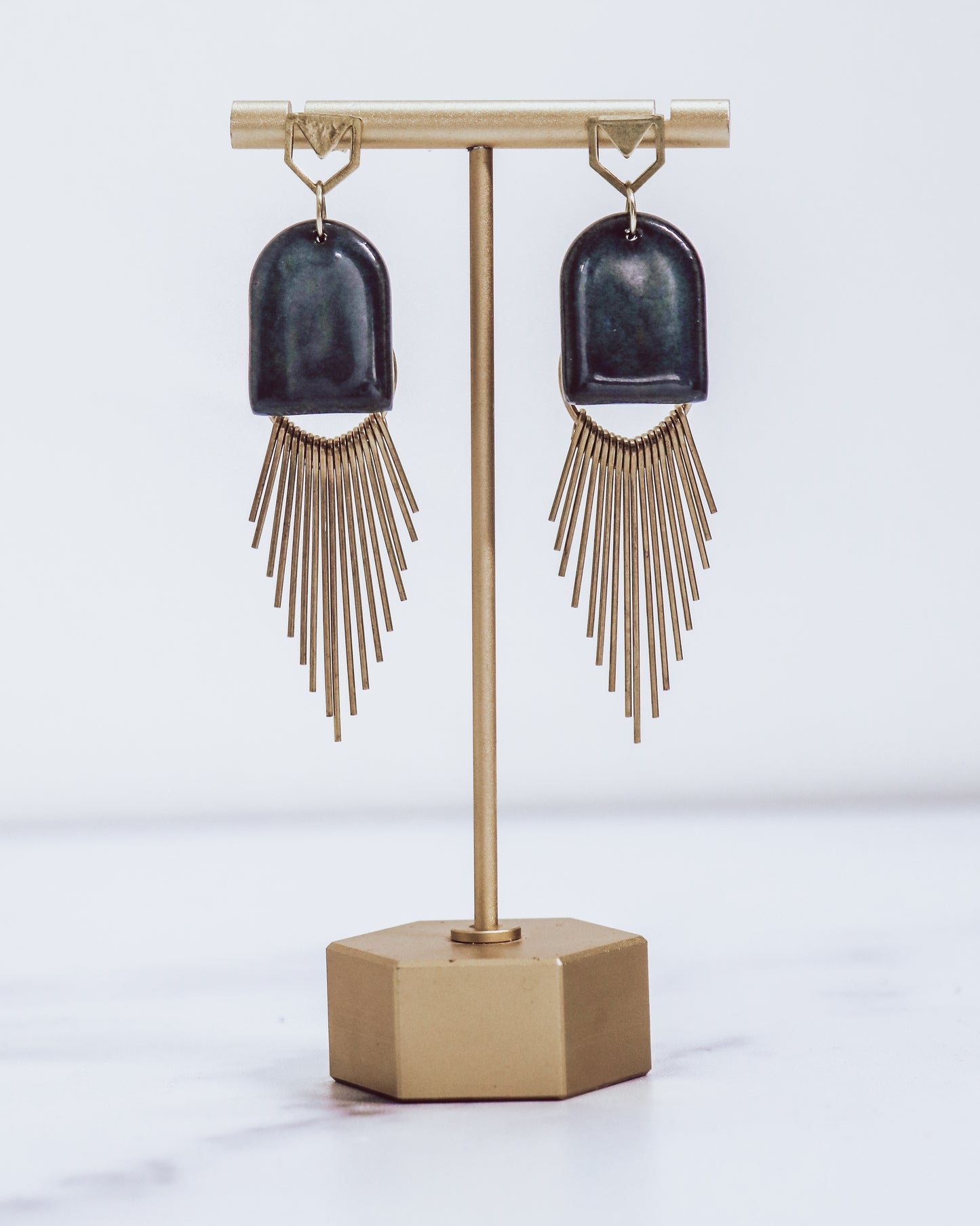 Ceramic and brass arch tassel earrings with gold fringe, shown in midnight blue on a gold stand.