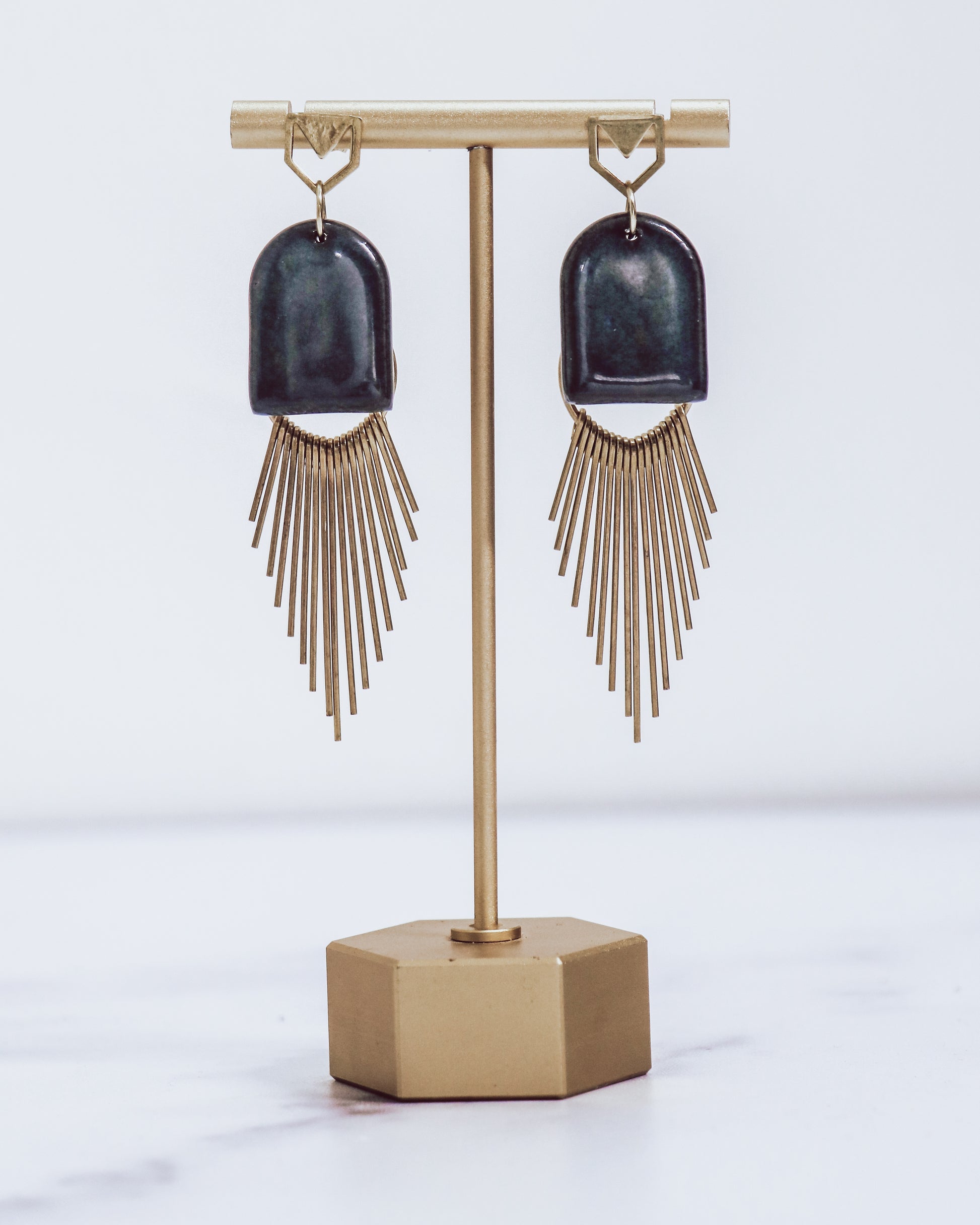 Ceramic and brass arch tassel earrings with gold fringe, shown in midnight blue on a gold stand.