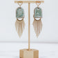Ceramic and brass arch tassel earrings with gold fringe, shown in pastel blue on a gold stand.