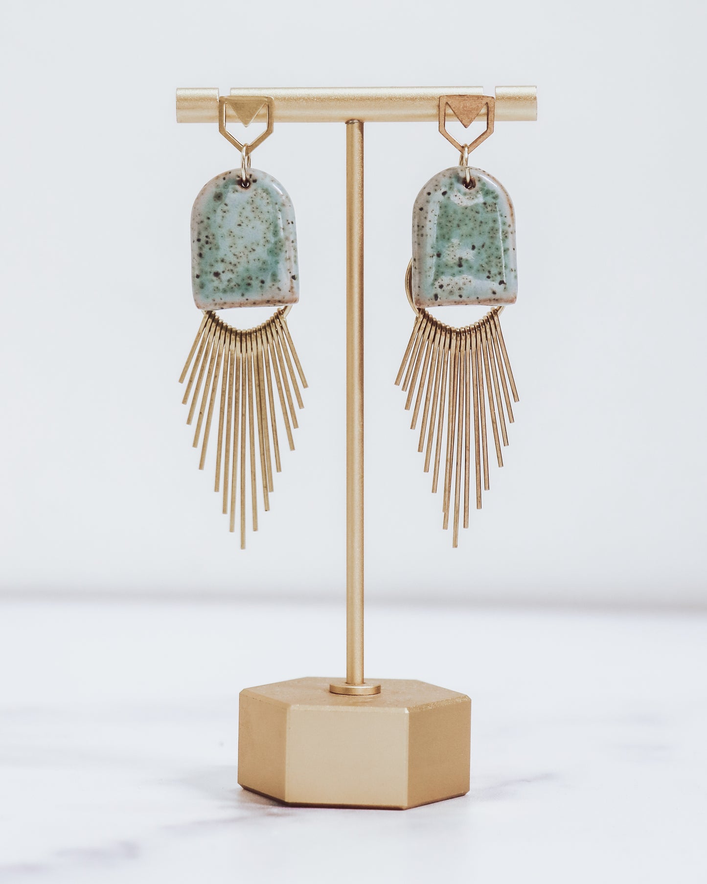 Ceramic and brass arch tassel earrings with gold fringe, shown in pastel blue on a gold stand.