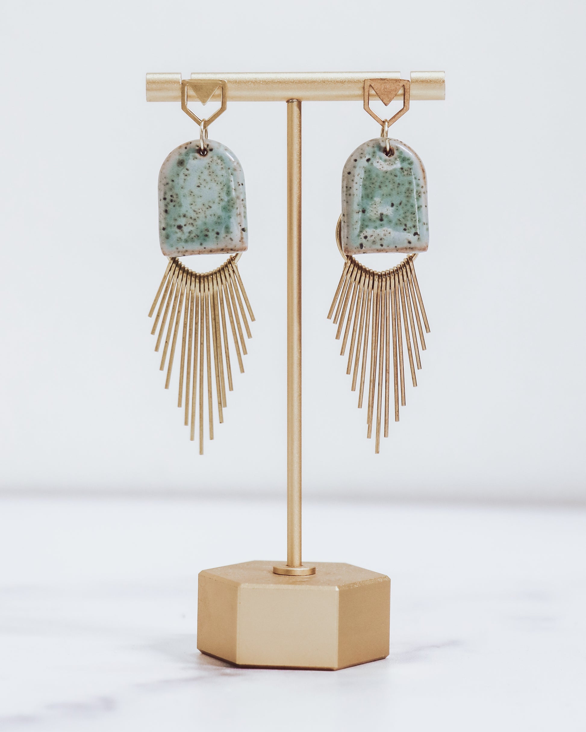 Ceramic and brass arch tassel earrings with gold fringe, shown in pastel blue on a gold stand.