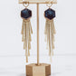 Long gold fringe tassel earrings with hexagon-shaped ceramic tops, shown in midnight blue on a gold stand. 