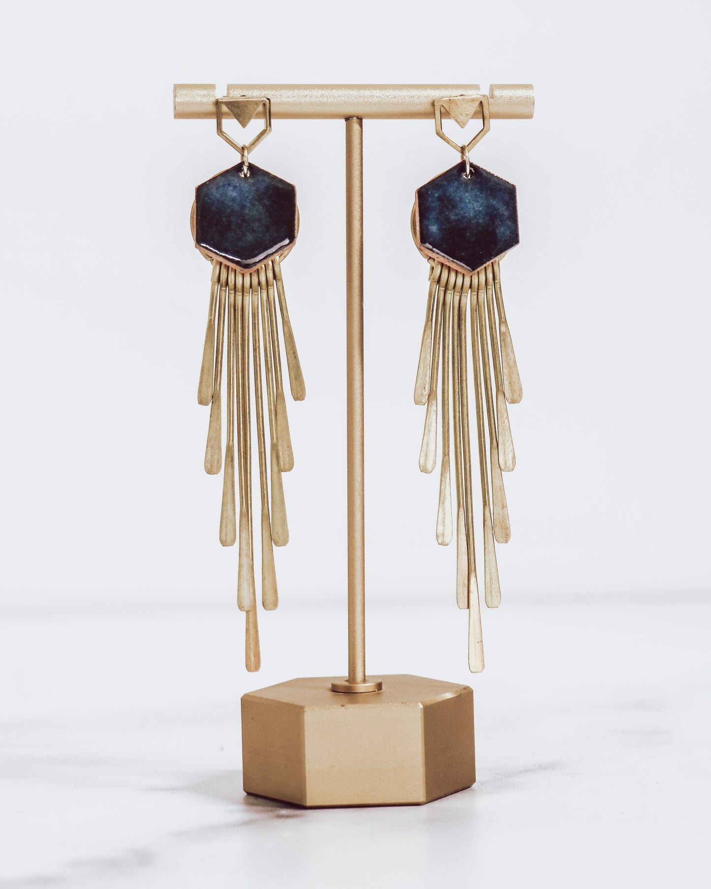 Long gold fringe tassel earrings with hexagon-shaped ceramic tops, shown in midnight blue on a gold stand. 