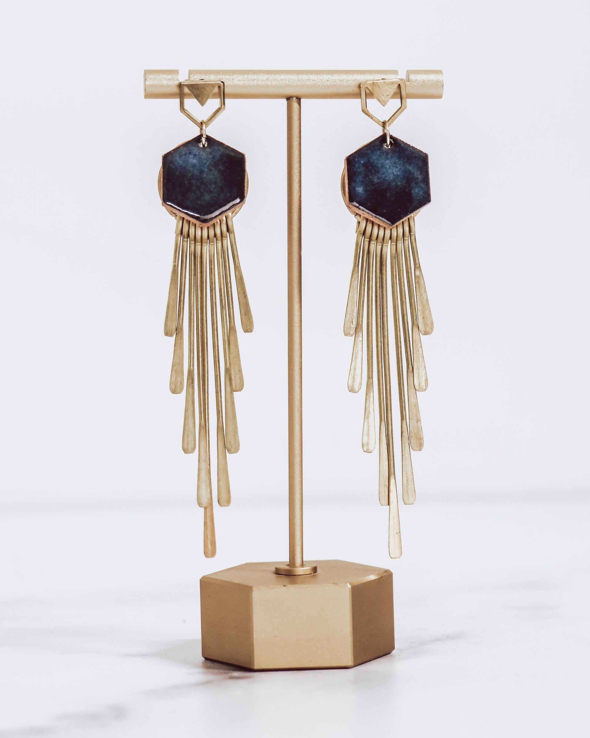 Long gold fringe tassel earrings with hexagon-shaped ceramic tops, shown in midnight blue on a gold stand. 