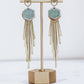 Long gold fringe tassel earrings with hexagon-shaped ceramic tops, shown in pastel blue on a gold stand. 