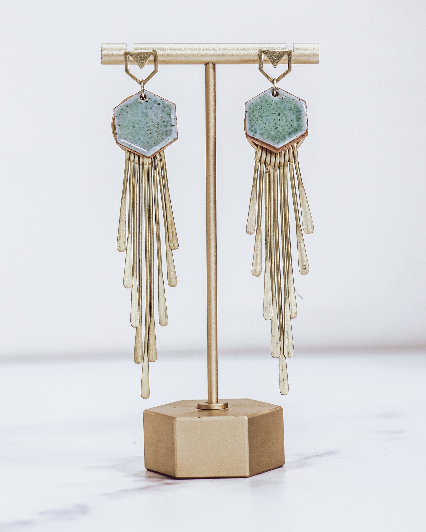 Long gold fringe tassel earrings with hexagon-shaped ceramic tops, shown in pastel blue on a gold stand. 
