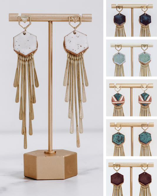 Long gold fringe tassel earrings with hexagon-shaped ceramic tops, shown in multiple glaze color options on a gold stand. 
