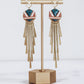 Long gold fringe tassel earrings with hexagon-shaped ceramic tops, shown in "nicola" glaze pattern  a gold stand.