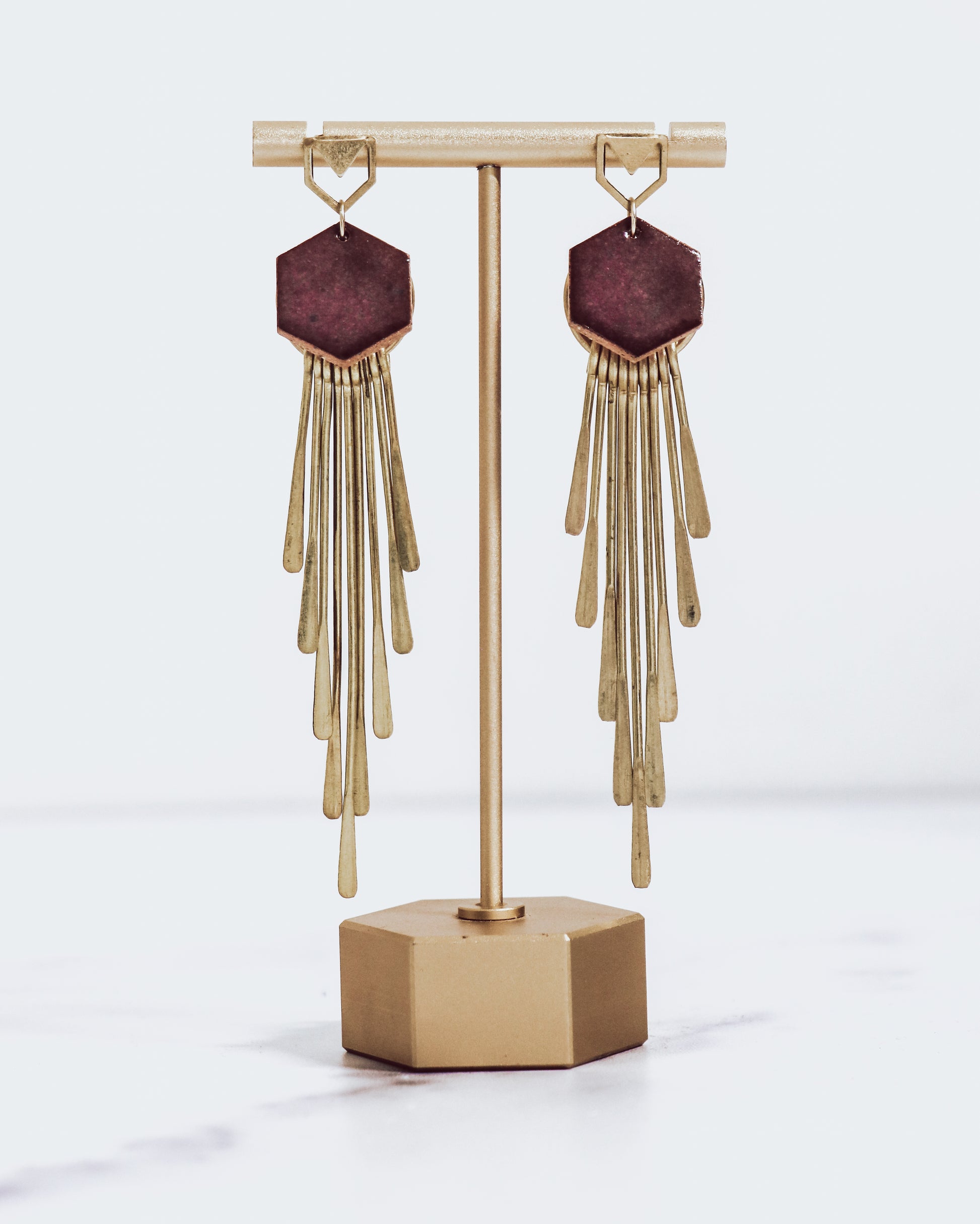 Long gold fringe tassel earrings with hexagon-shaped ceramic tops, shown in burgundy on a gold stand.