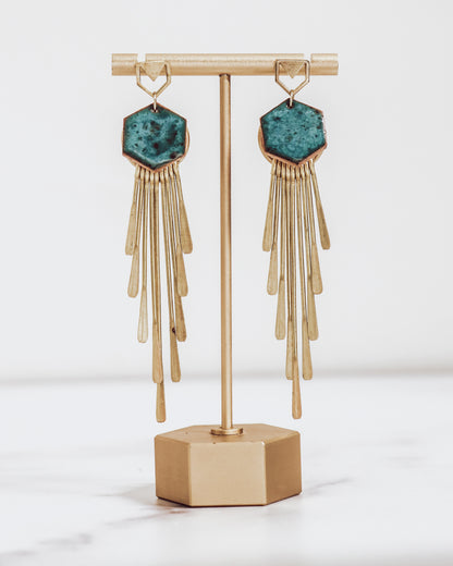Long gold fringe tassel earrings with hexagon-shaped ceramic tops, shown in turquoise on a gold stand.
