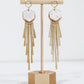 Long gold fringe tassel earrings with hexagon-shaped ceramic tops, shown in white on a gold stand.