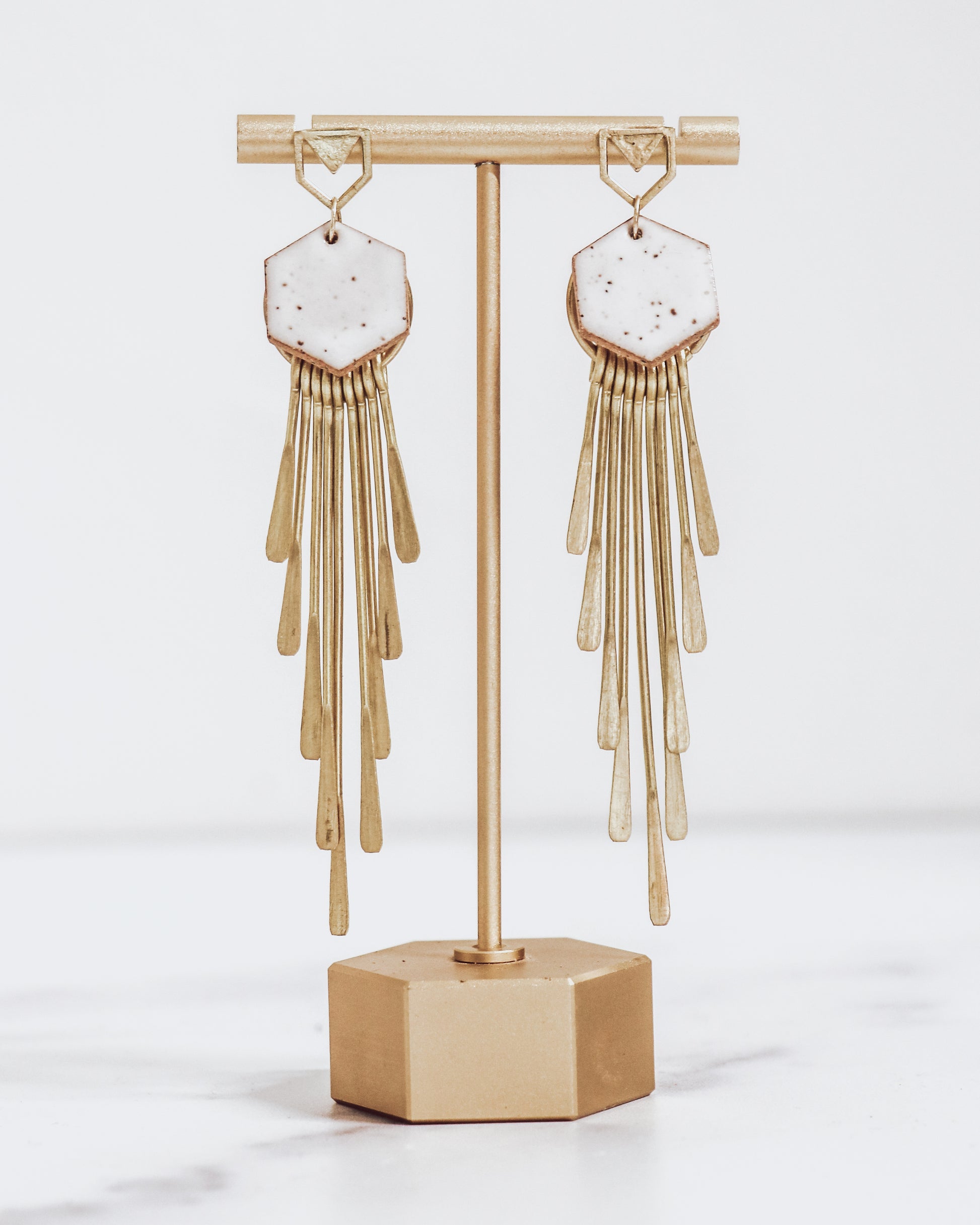 Long gold fringe tassel earrings with hexagon-shaped ceramic tops, shown in white on a gold stand.