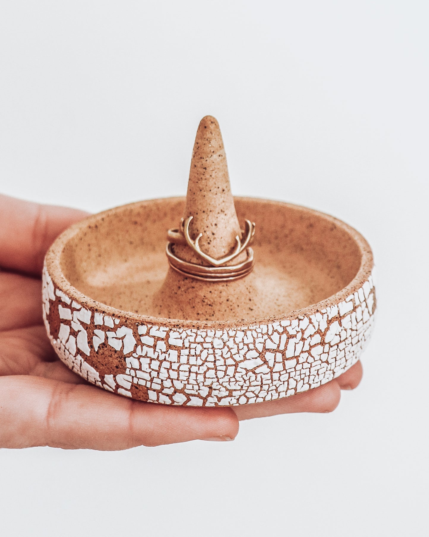 Ceramic ring holder with center cone, crackle white exterior, and speckled tan interior, shown in hand with rings for scale.