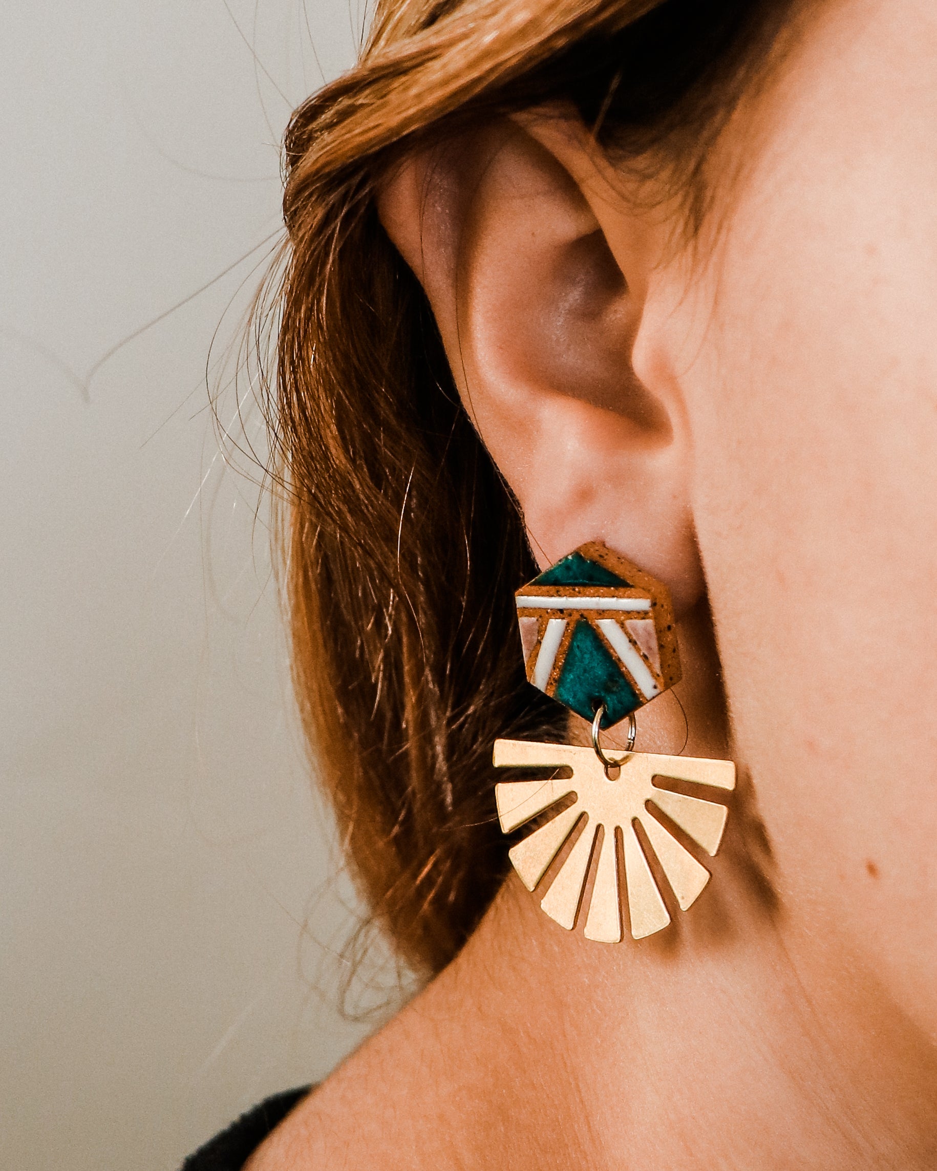 Pair of handmade geometric ceramic and brass earrings modeled on a young woman, featuring teal glaze, white accents, and fan shaped brass drops. 