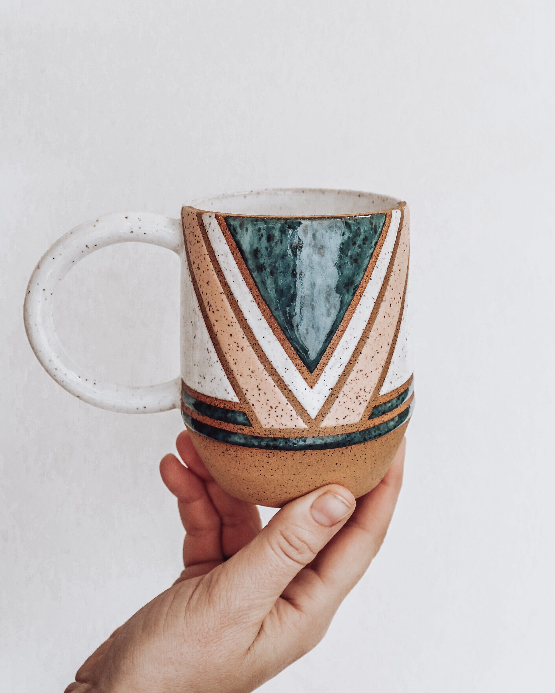 A ceramic mug with a geometric pattern in shades of green pink and white held in a person's hand.