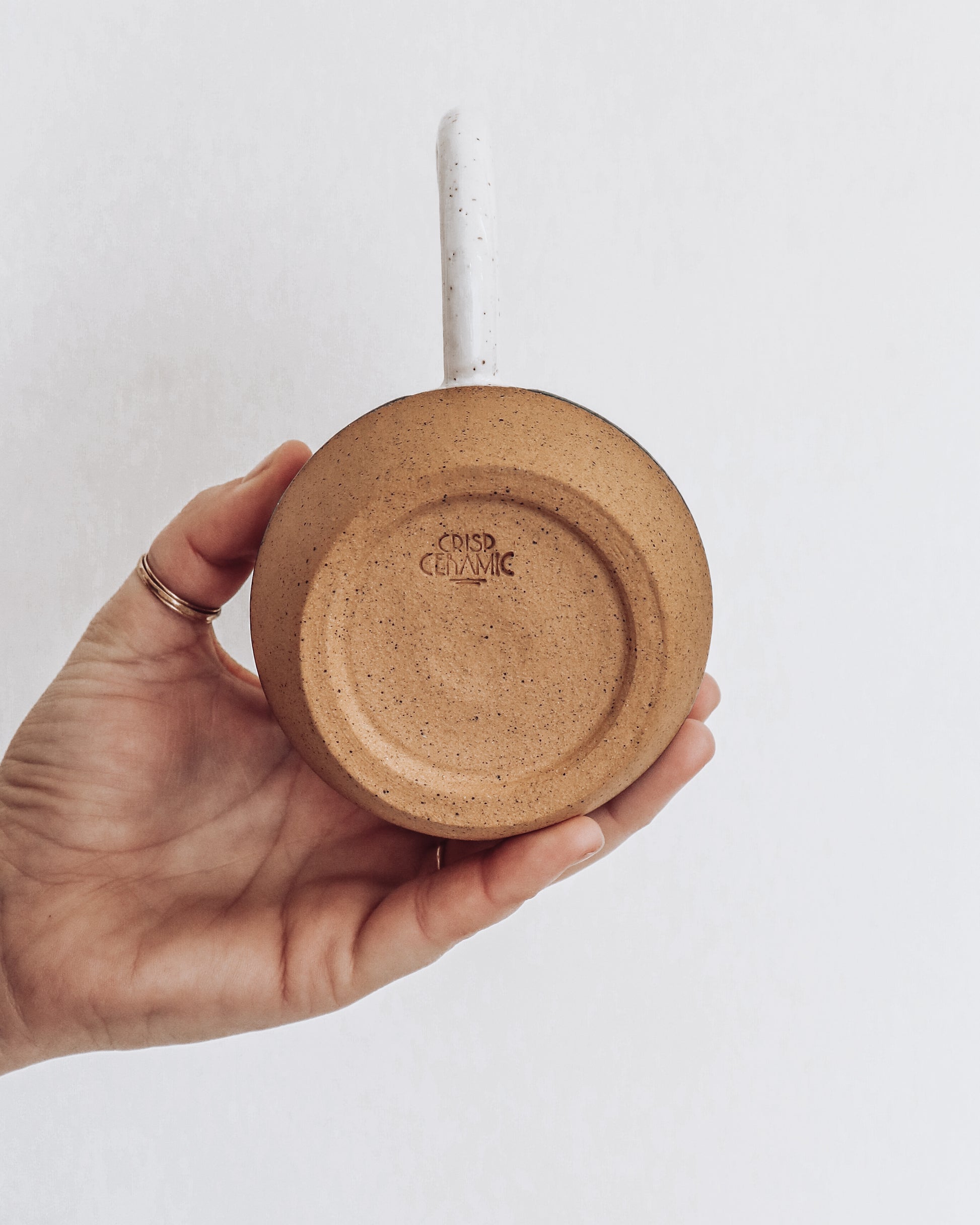 A ceramic mug with a geometric pattern in shades of green pink and white held in a person's hand. view show unglazed bottom of mug with crisp ceramic makers mark.