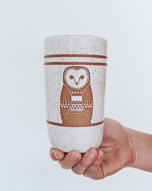 A ceramic tumbler showing geometric owl design with glossy white glaze, held in a person's hand against a white background.