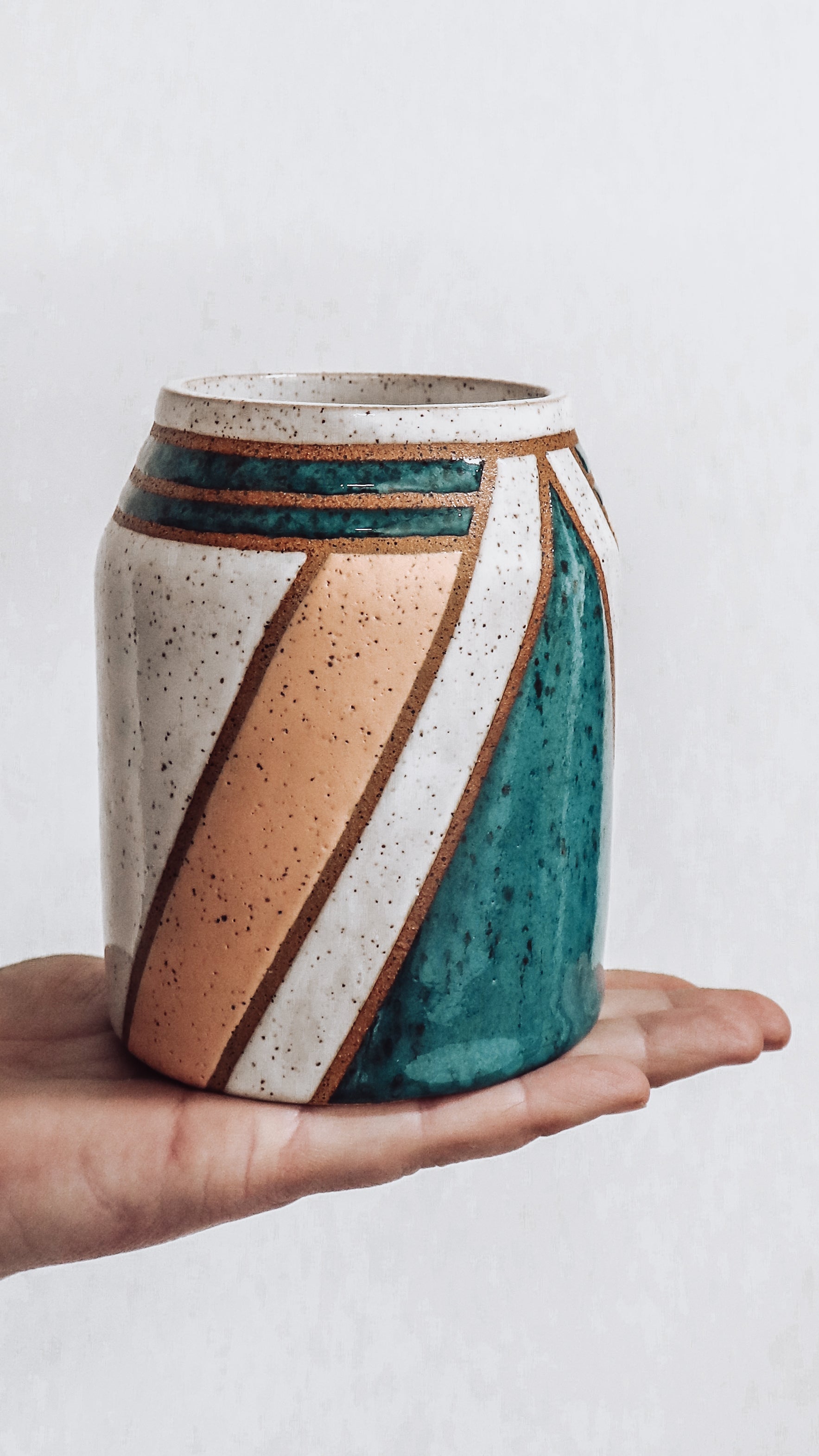 hand holding a handmade ceramic vase featuring teal, white and pink glaze with geometric pattern.