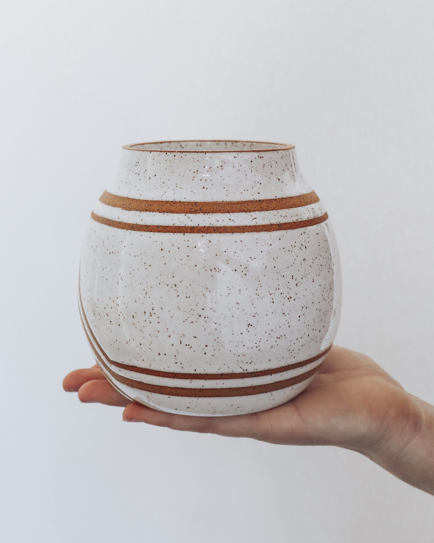  Hand holding a handmade speckled ceramic vase with geometric stripe detailing in warm brown tones. 