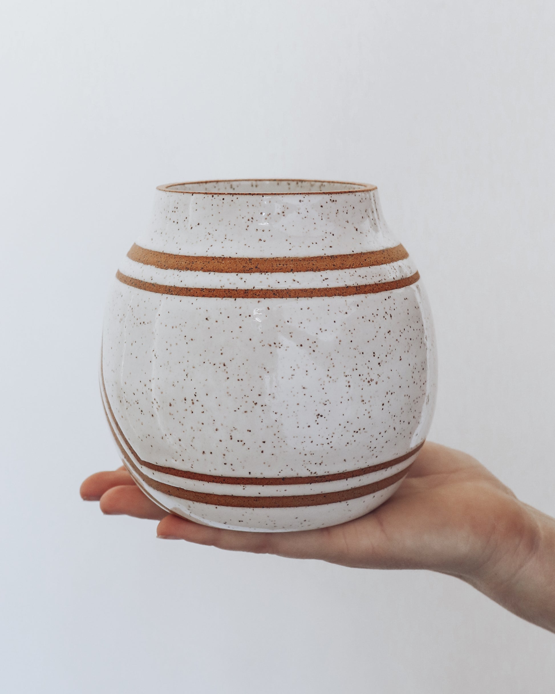  Hand holding a handmade speckled ceramic vase with geometric stripe detailing in warm brown tones. 