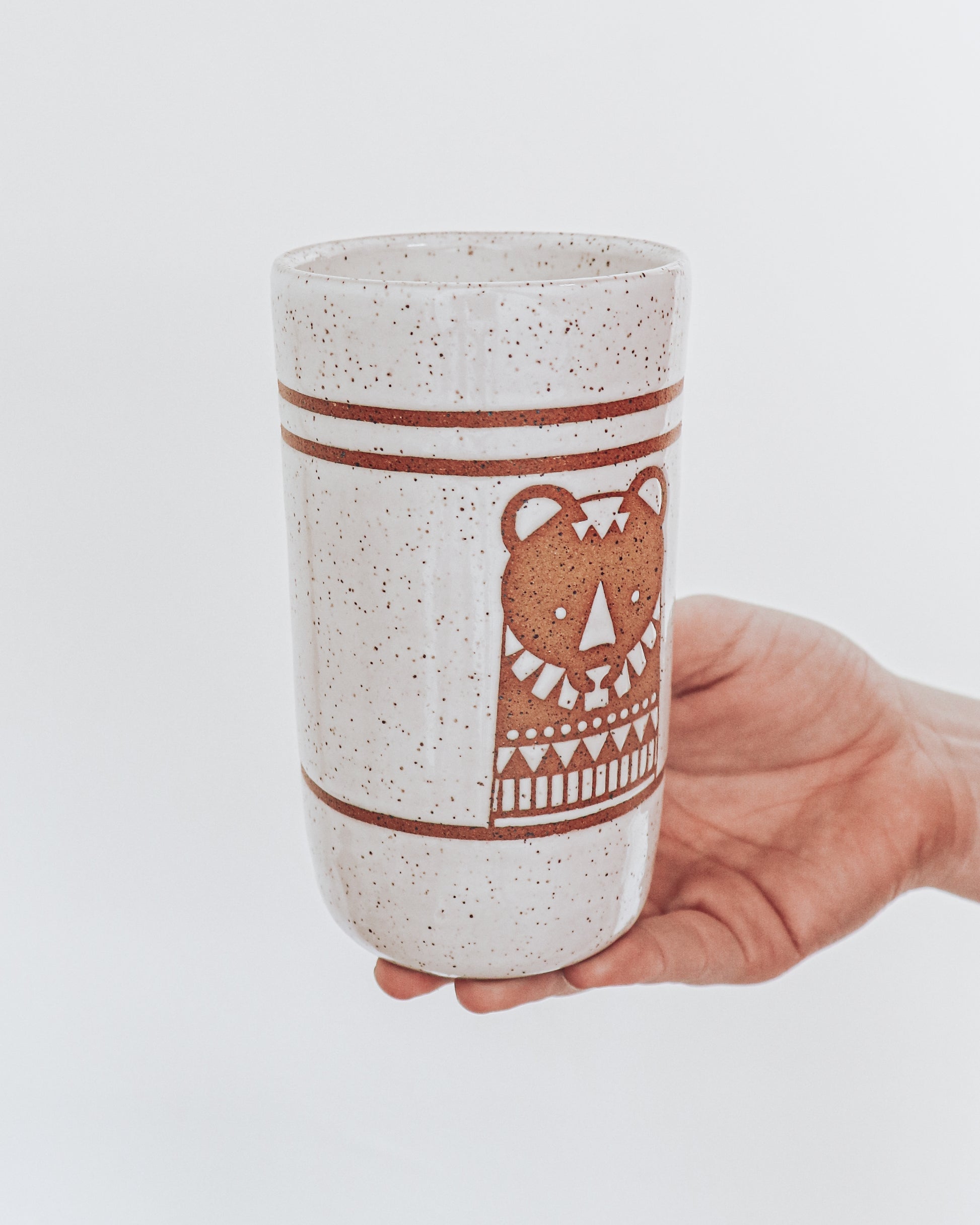 Hand Holding a handmade speckled ceramic tumbler featuring a brown illustrated bear design with geometric detailing. 
