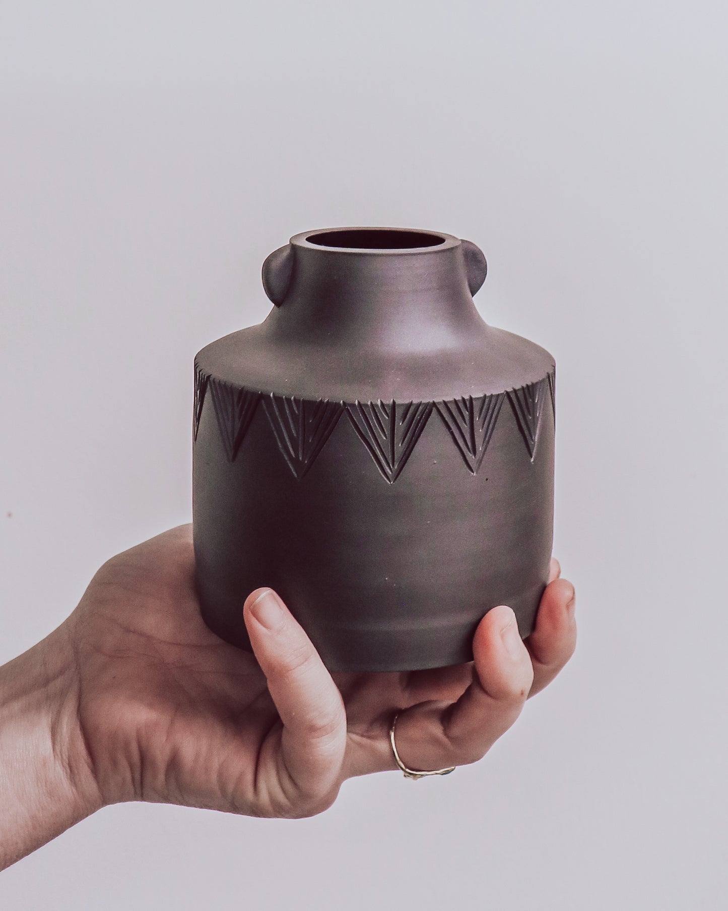 hand holding dark matte ceramic vase with carved geometric detailing and small sculpted side details.