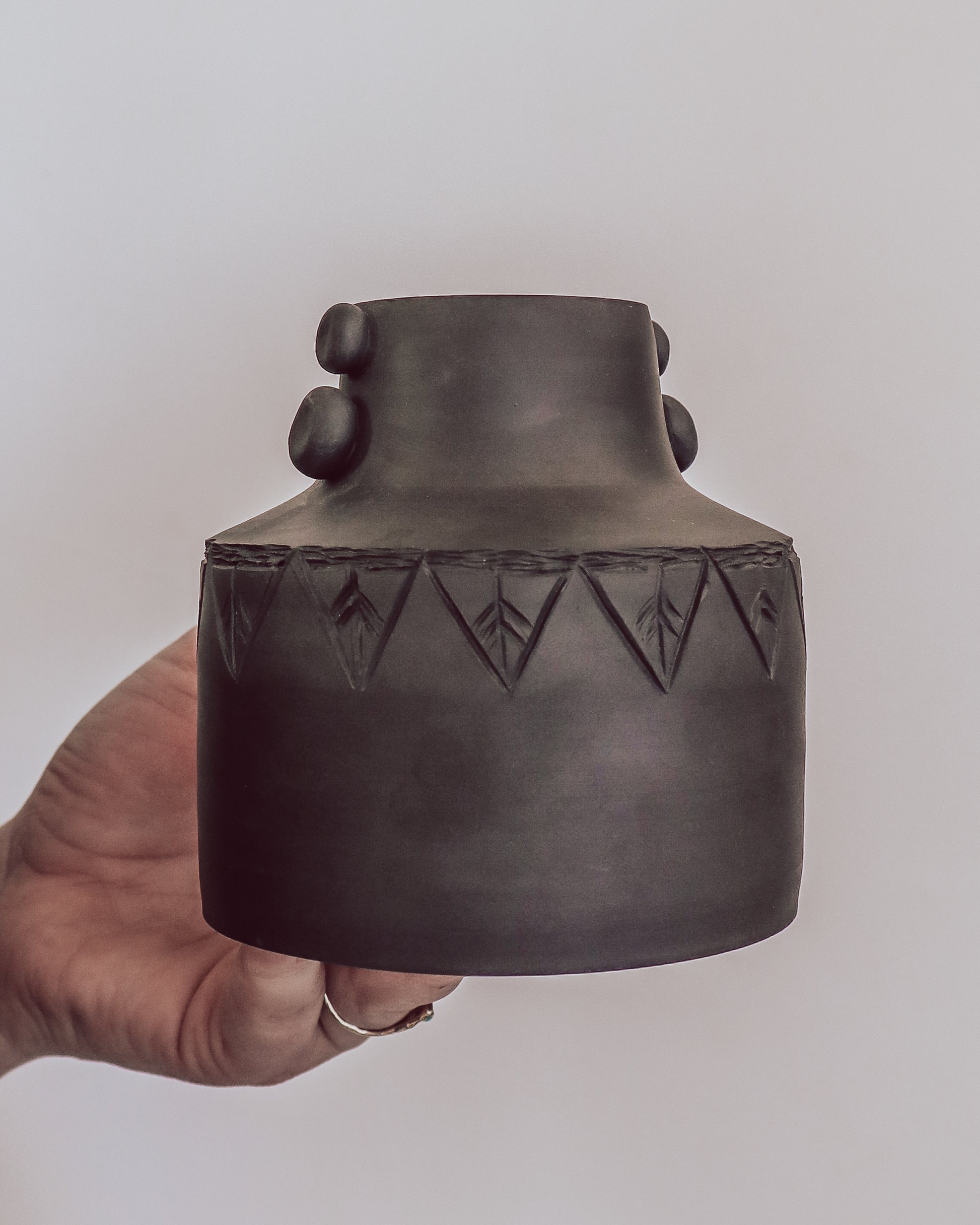 hand holding dark matte ceramic vase with carved geometric detailing and small sculpted side details.