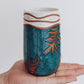 Handmade ceramic tumbler in deep teal glaze with leaf design and speckled white rim, shown held in hand for scale.
