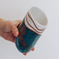 Handmade ceramic tumbler in deep teal glaze with leaf design and speckled white rim, shown held in hand for scale.