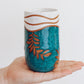 Handmade ceramic tumbler in deep teal glaze with leaf design and speckled white rim, shown held in hand for scale.