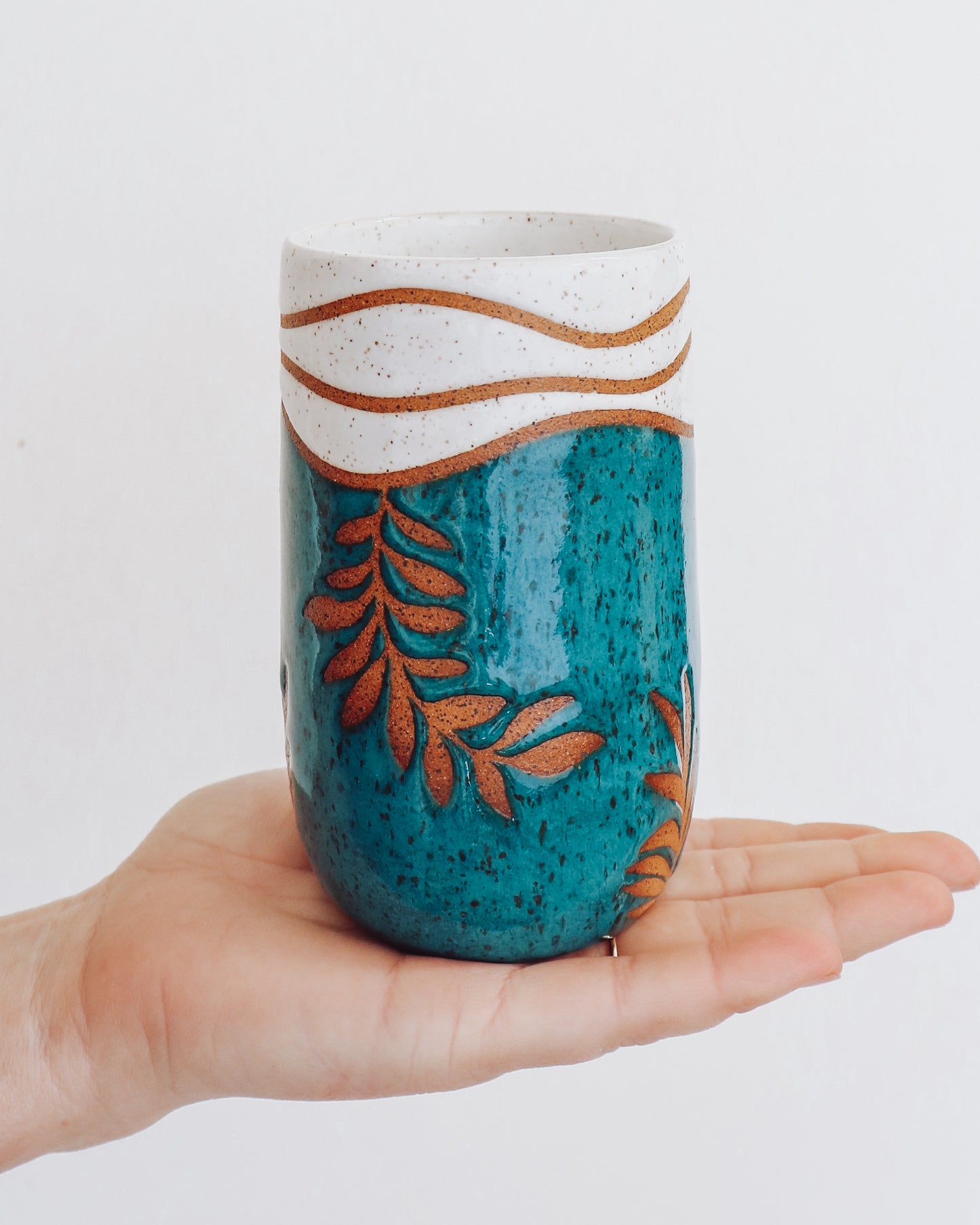 Handmade ceramic tumbler in deep teal glaze with leaf design and speckled white rim, shown held in hand for scale.