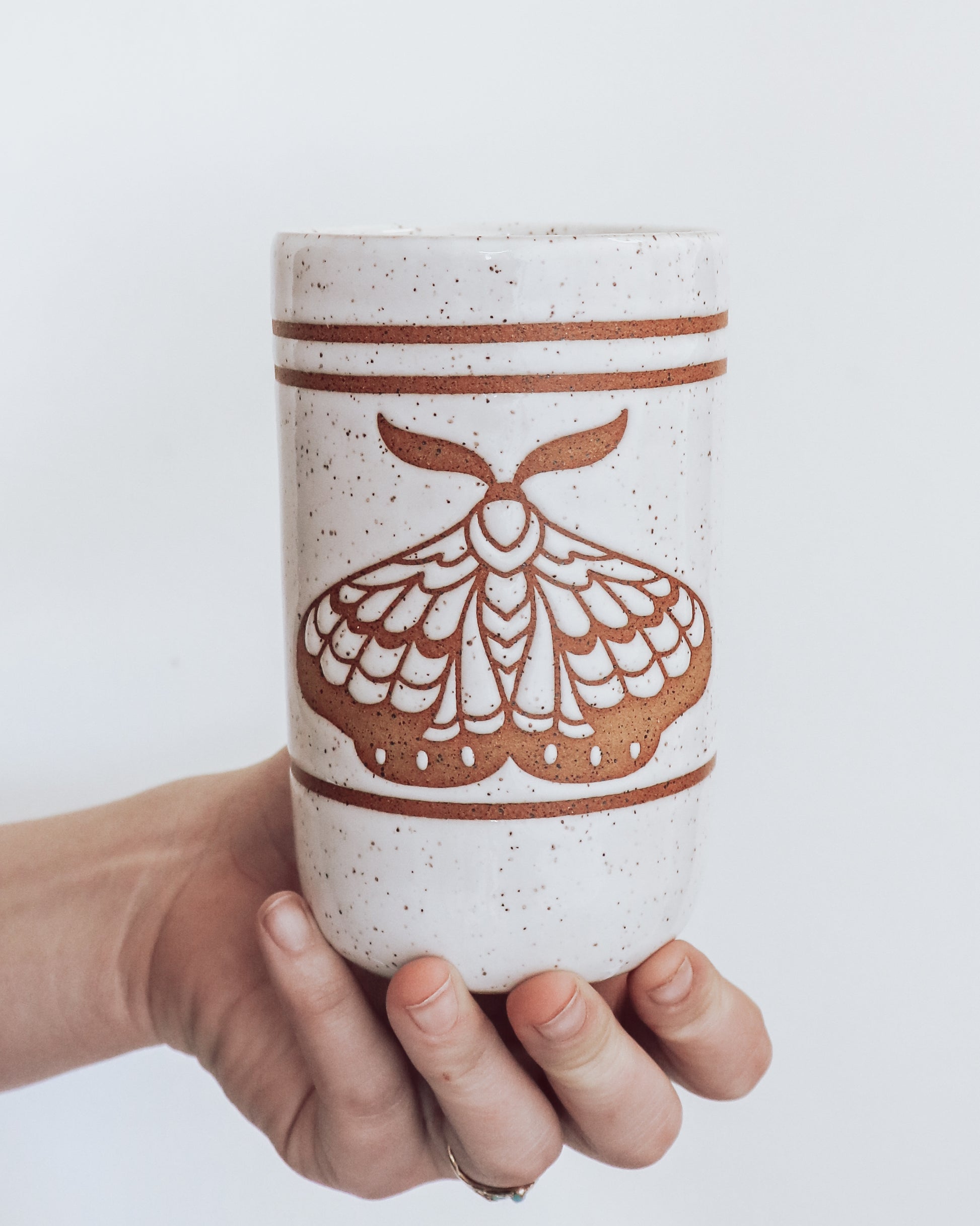 A ceramic tumbler showing geometric moth design with glossy white glaze, held in a person's hand against a white background.