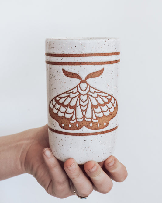 A ceramic tumbler showing geometric moth design with glossy white glaze, held in a person's hand against a white background.