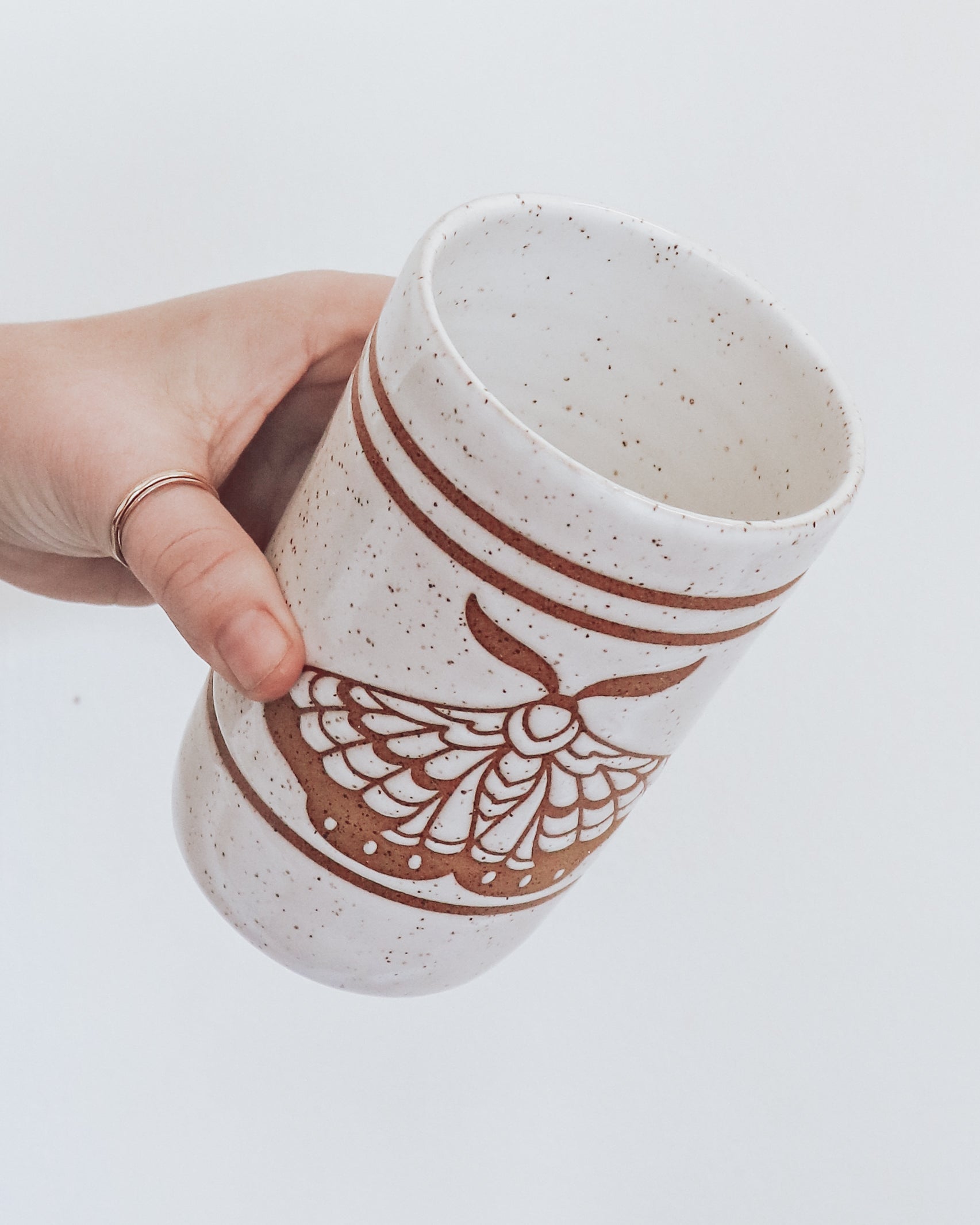 A ceramic tumbler showing geometric moth design with glossy white glaze, held in a person's hand against a white background.
