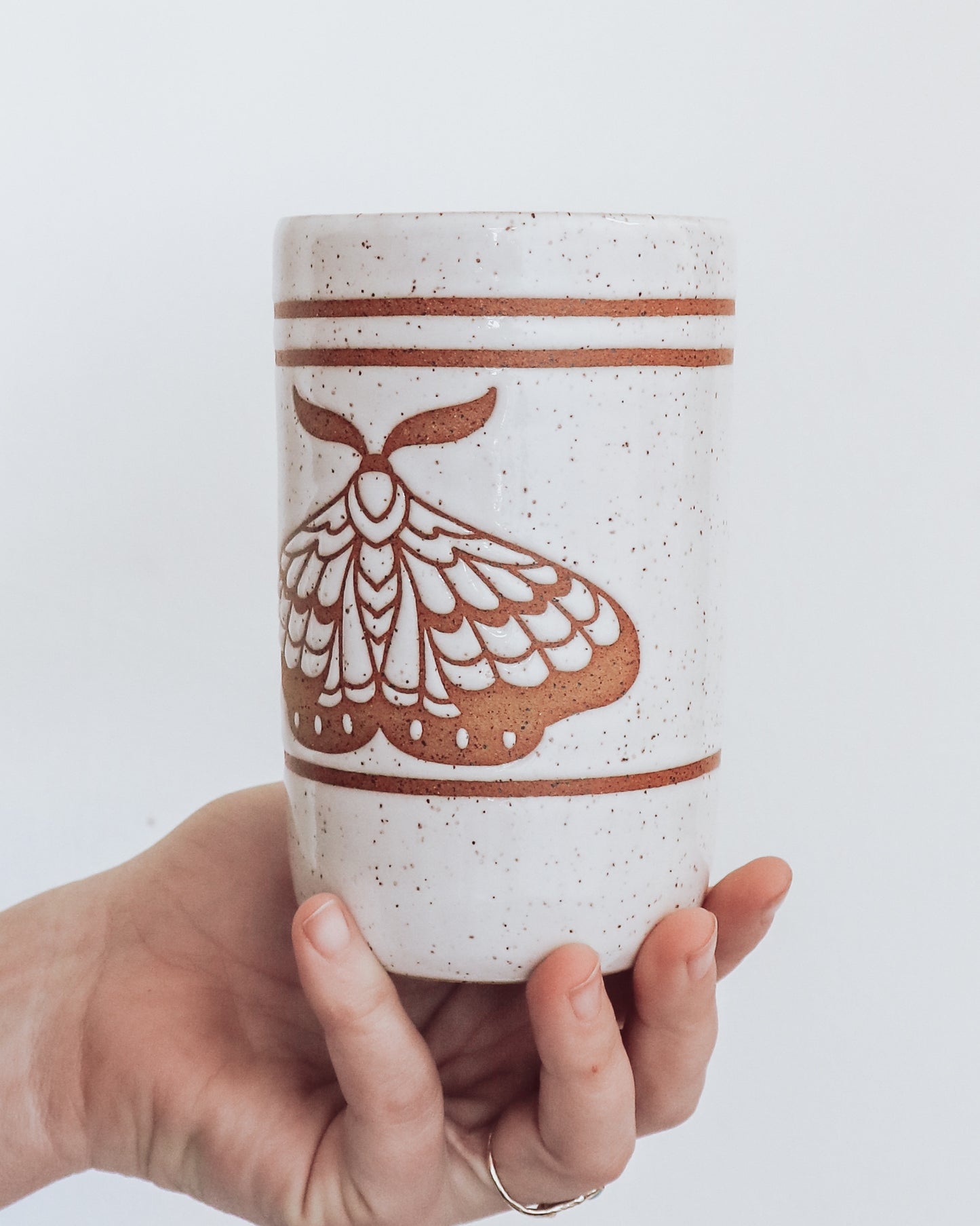 A ceramic tumbler showing geometric moth design with glossy white glaze, held in a person's hand against a white background.
