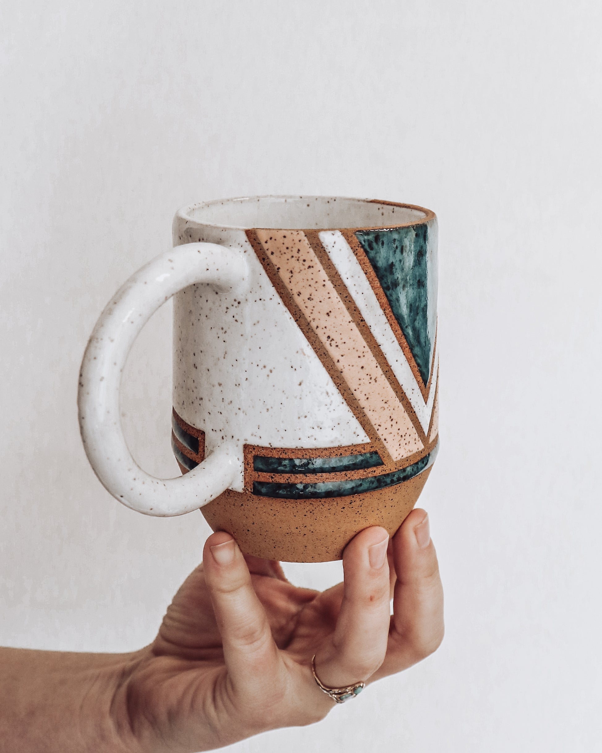 A ceramic mug with a geometric pattern in shades of green pink and white held in a person's hand.