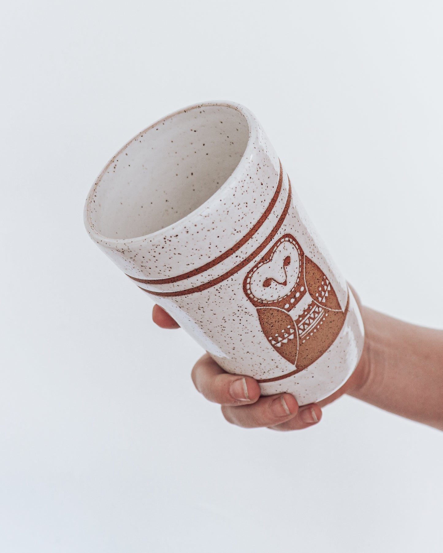 A ceramic tumbler showing geometric moth design with glossy white glaze, held in a person's hand against a white background.
