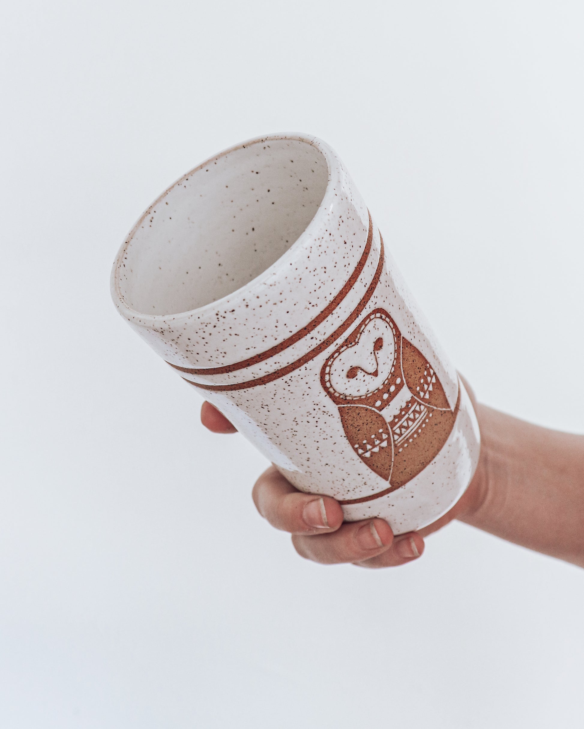 A ceramic tumbler showing geometric moth design with glossy white glaze, held in a person's hand against a white background.