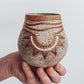 A hand holding a small brown and beige ceramic vase with a crackled texture and geometric patterns.