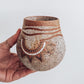 A hand holding a small brown and beige ceramic vase with a crackled texture and geometric patterns.