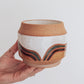 Hand holding a handmade ceramic planter with white glaze and wavy multicolored stripe detailing. 