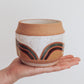 Hand holding a handmade ceramic planter with white glaze and wavy multicolored stripe detailing. 
