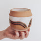 Hand holding a handmade ceramic planter with white glaze and wavy multicolored stripe detailing. 