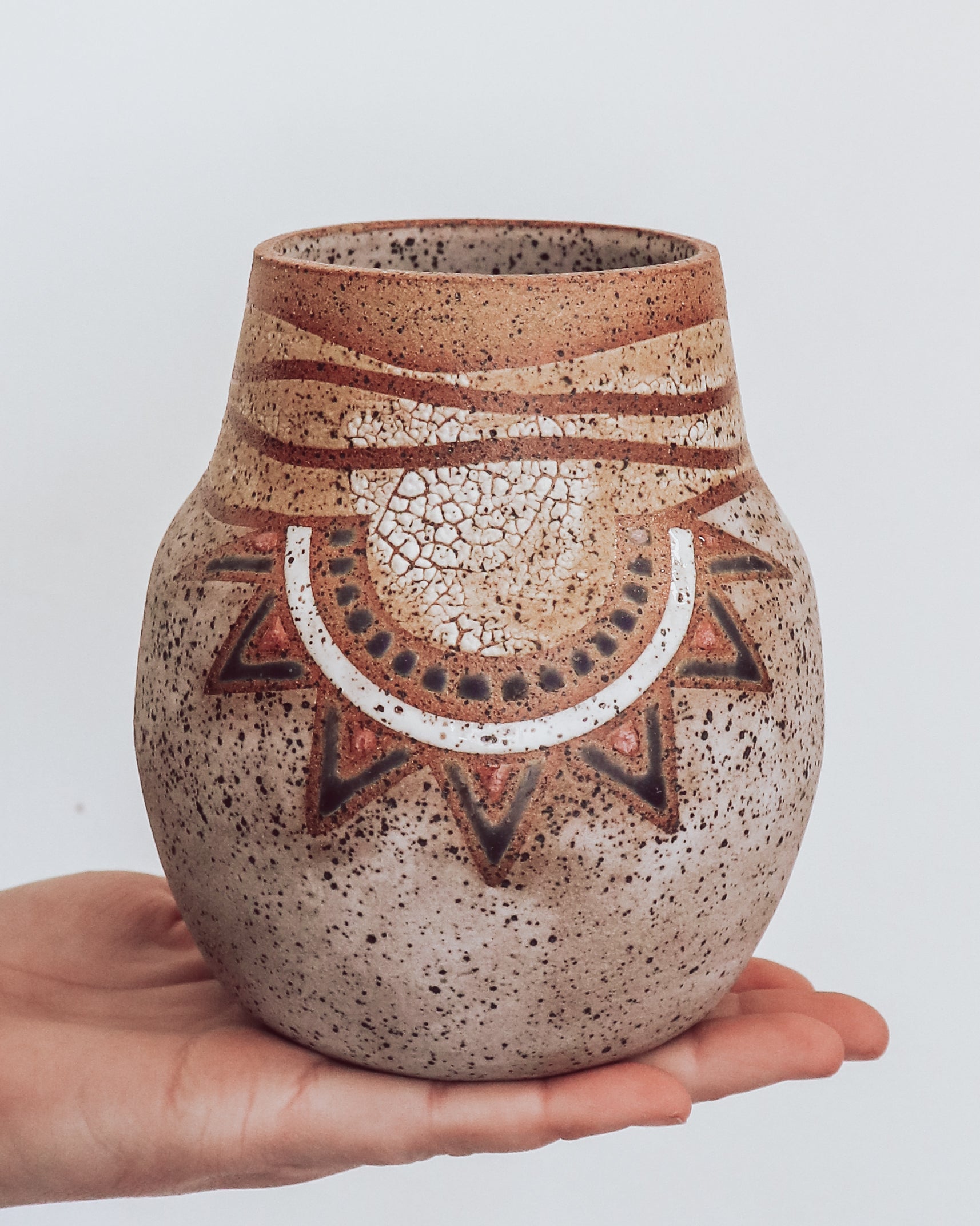 A hand holding a small brown and beige ceramic vase with a crackled texture and geometric patterns.